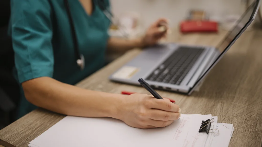 Professionista sanitario in divisa che prende appunti su un foglio accanto a un computer portatile